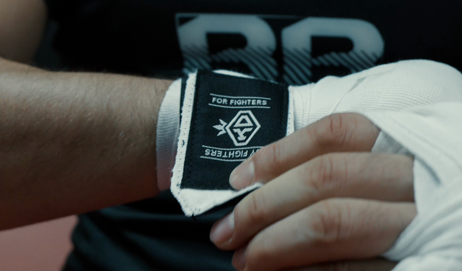 KOStyle Hand Wraps. An athlete secures a white boxing hand wrap on their wrist with a black velcro strap featuring the text "FOR FIGHTERS BY FIGHTERS" and a logo, while wearing a dark athletic shirt with a white claw mark graphic.